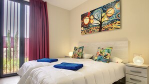 Quarto com cama grande, duas almofadas, mesa de cabeceira, pintura e janela com cortinas.
