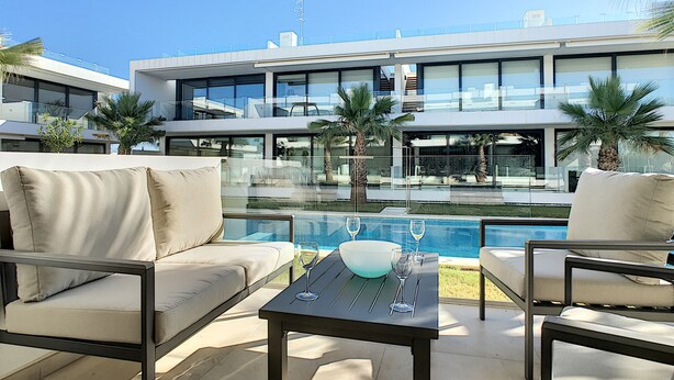 Uma área de estar moderna ao ar livre com piscina, destacando uma mesa de vidro com taças de vinho e uma tigela, cercada por sofás e cadeiras.