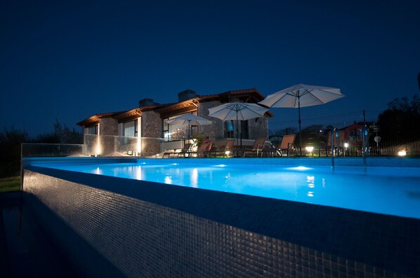 A swimming pool with lounge chairs and umbrellas, a stone building with a tiled roof, and a clear night sky.