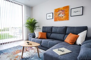 A modern living room with a grey sofa, orange cushions, a round wooden coffee table, and a large window with blinds. -  With pool and views of the jesters. Ideal for families (LLANES)