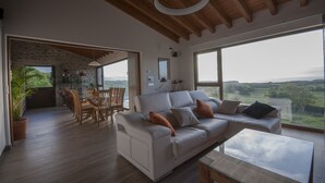 Un vaste espace de vie comprenant un grand canapé blanc, une table à manger en bois et des chaises. Une grande fenêtre offre une vue sur la campagne.