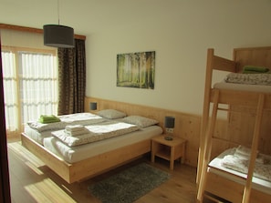 A bedroom with a wooden bunk bed, a single bed with floral bedding, a small bedside table with a lamp, a painting of a forest, and a window with curtains. - Perfect View with large balcony overlooking Lienz, swimming pool, sauna, parking (Lienz)