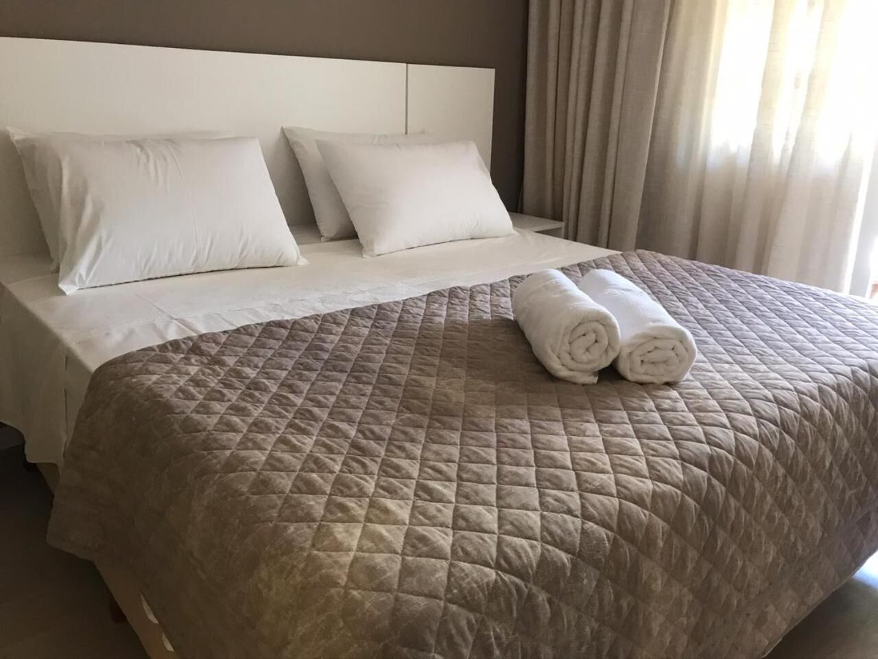A neatly made bed with a brown quilt, white pillows, and rolled white towels.
