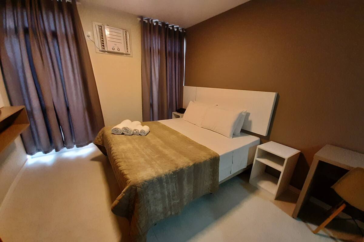 A hotel room with a bed, a bedside table, a chair, a desk, and curtains.