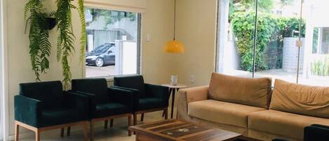 Una sala moderna con un sofá color beige, dos sillones verde oscuro, una mesa de centro de madera y plantas colgantes.
