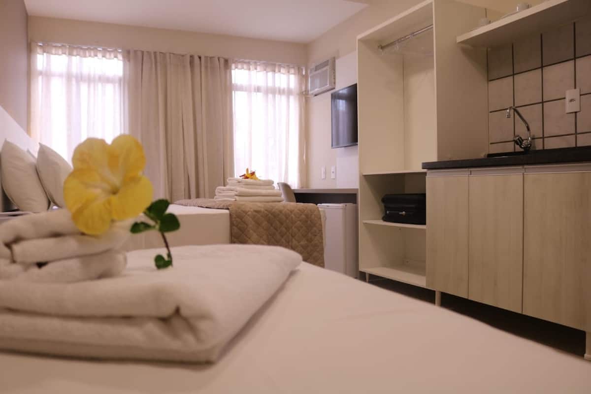 A hotel room with a bed, a small table with towels, a window with curtains, a TV mounted on the wall, and a kitchenette area.