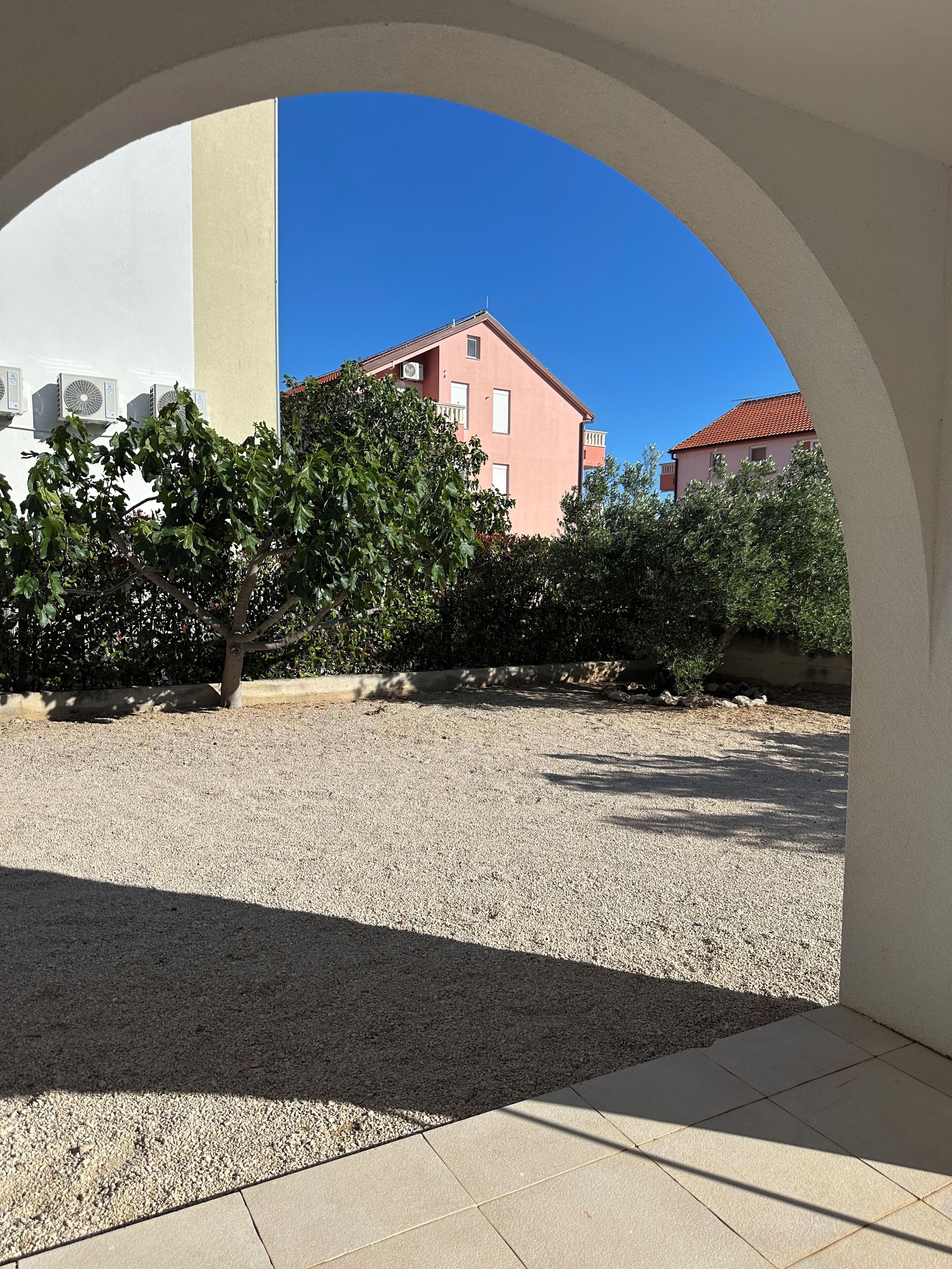 Un patio exterior con suelo de grava, un edificio blanco con unidades de aire acondicionado y un edificio rosa al fondo.