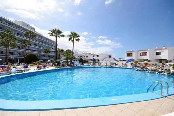 A swimming pool with lounge chairs and umbrellas, surrounded by palm trees and buildings. - Club Atlantis 89 - Two Bed (Costa Adeje)