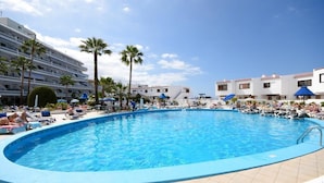 Une grande piscine extérieure entourée de chaises longues et de parasols, avec des palmiers et un immeuble à plusieurs étages en arrière-plan.