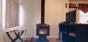A room with a wood-burning stove, a television on a stand, and a window with blinds. - Cozy cottage in  Warwick's West Bay (Warwick)