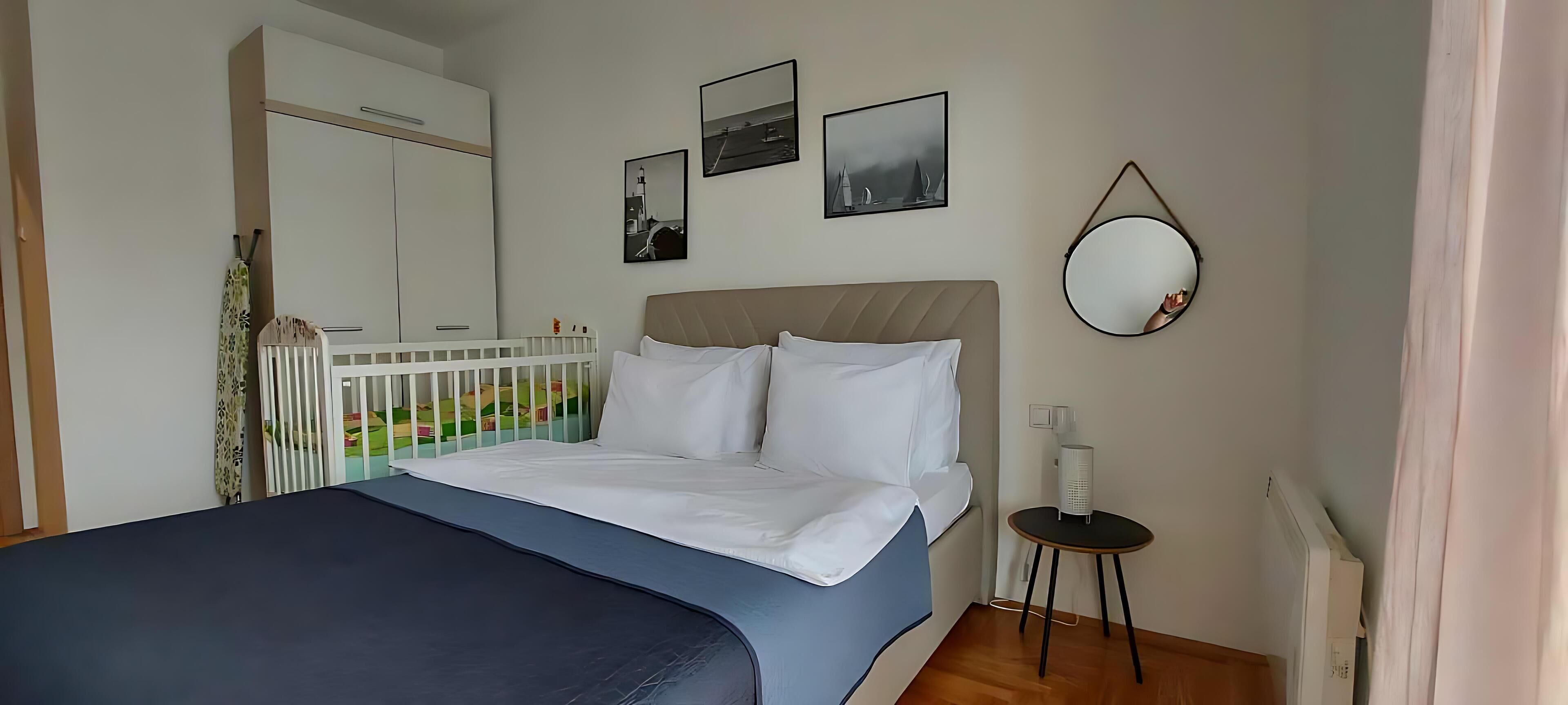 A bedroom with a bed, a crib, a round mirror, and a small table.