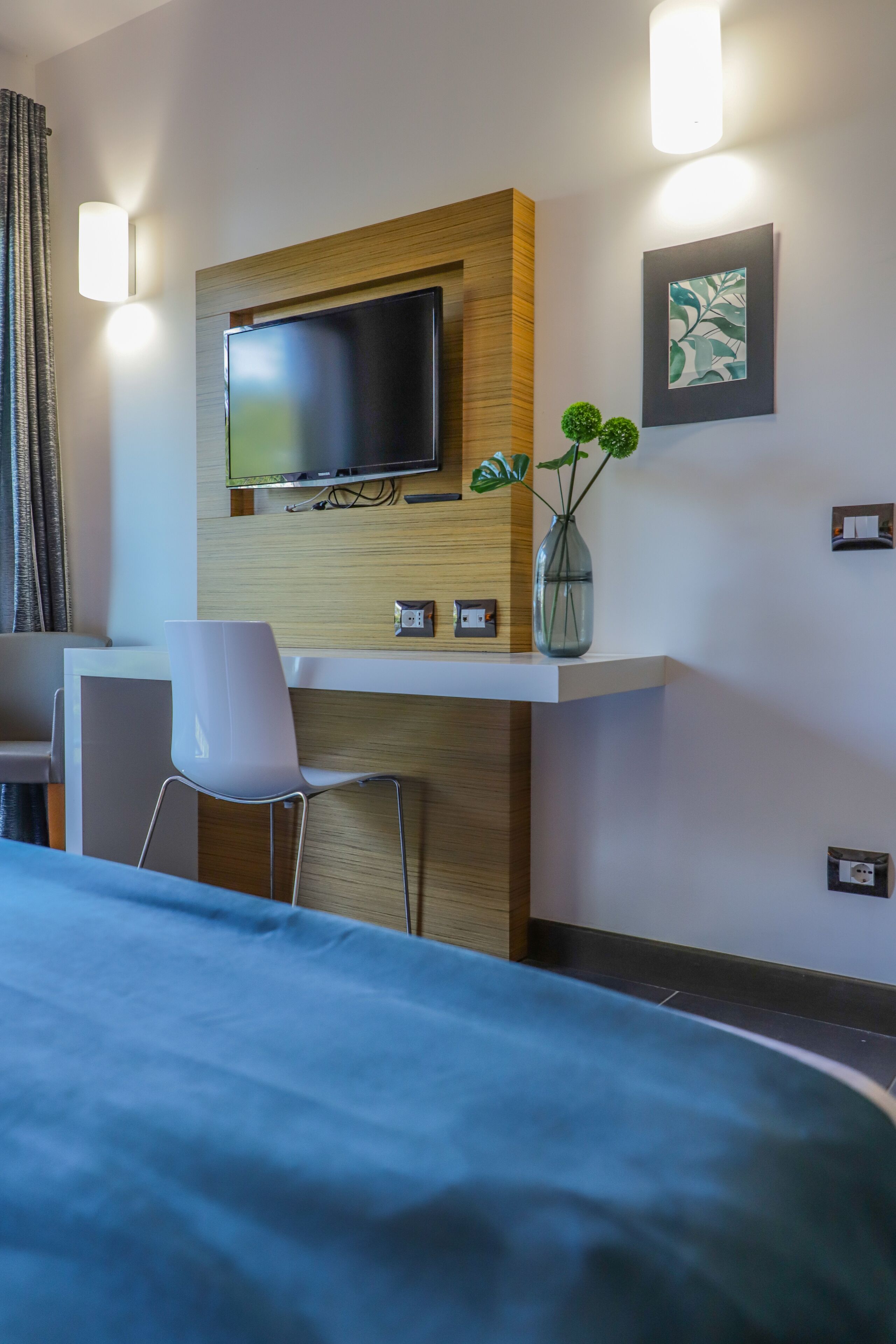 A hotel room with a bed, a desk with a chair, a flat-screen TV mounted on a wooden panel, a vase with plants, and a framed artwork on the wall.
