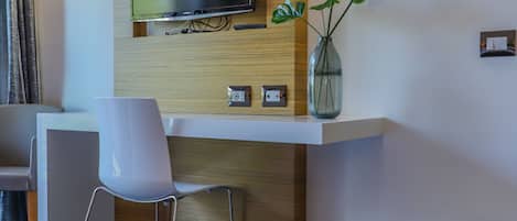 A hotel room with a bed, a desk with a chair, a flat-screen TV mounted on a wooden panel, a vase with plants, and a framed artwork on the wall.