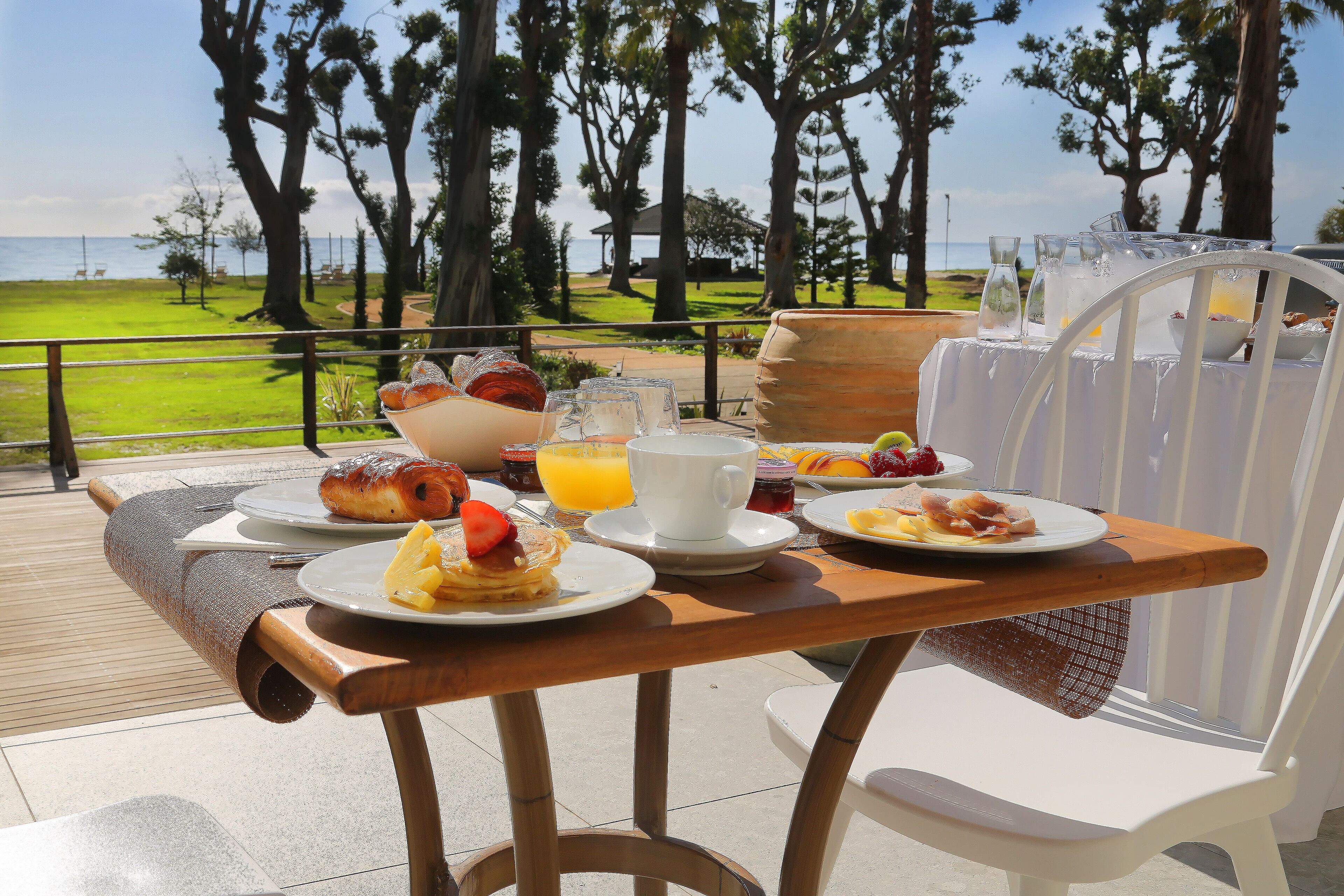 Mesa para comer al aire libre con alimentos para el desayuno, vista al océano y árboles al fondo.