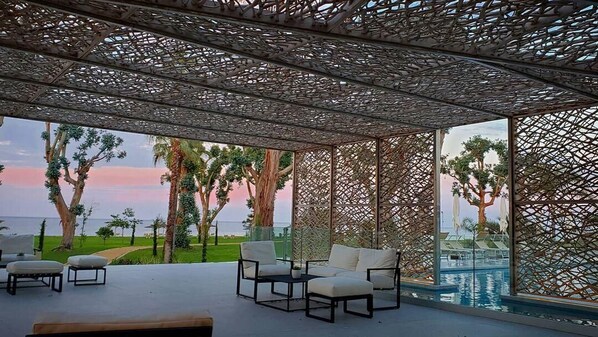 Un área de estar al aire libre con techo de celosía y mobiliario exterior, con vista a la piscina.