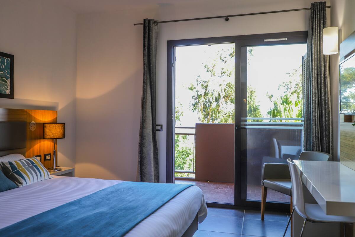 Une chambre d’hôtel avec un lit, un bureau et un balcon donnant sur un espace verdoyant.