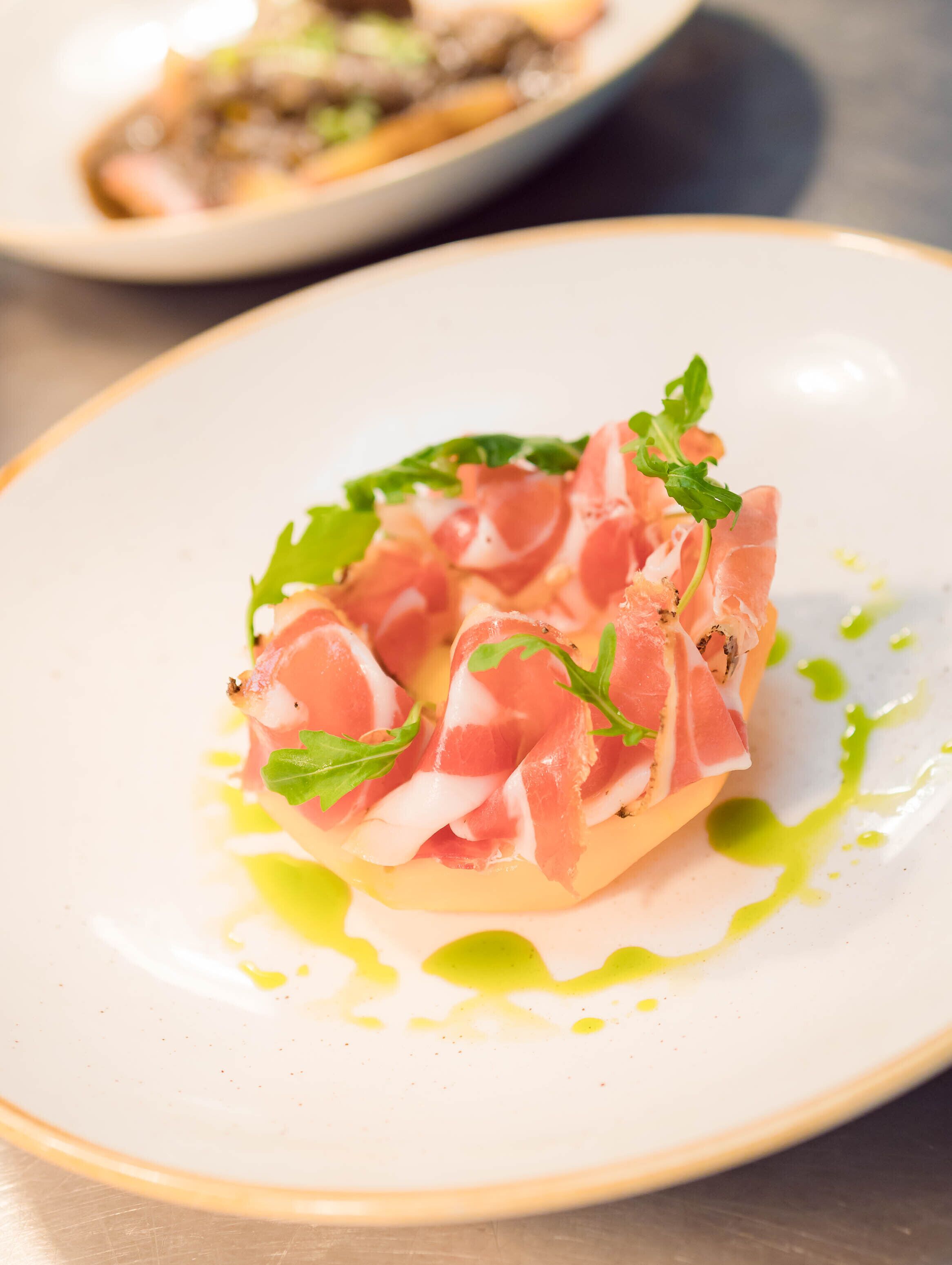 Een bord met een gerecht bestaande uit dun gesneden vlees, mogelijk parmaham, versierd met groene blaadjes en een lichtkleurige saus.