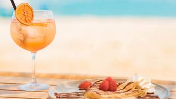 A glass of iced drink with a slice of orange on a beach with a plate of waffles and raspberries.