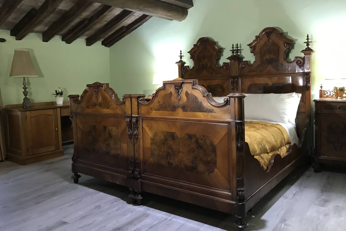 Een traditioneel houten bed met een hoge hoofdbord, een nachtkastje met een lamp en een houten dressoir in een kamer met een schuine kap.