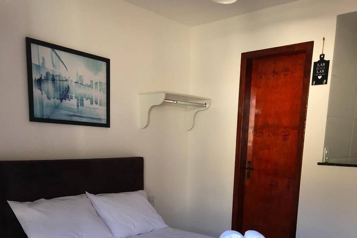 A neatly made bed with white and red linens, a framed cityscape picture on the wall, and a wooden door with a small sign.