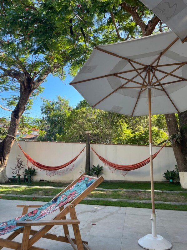 Uma cadeira rede sob um guarda-sol branco, um cavalete de madeira e um jardim com árvores e uma parede branca.