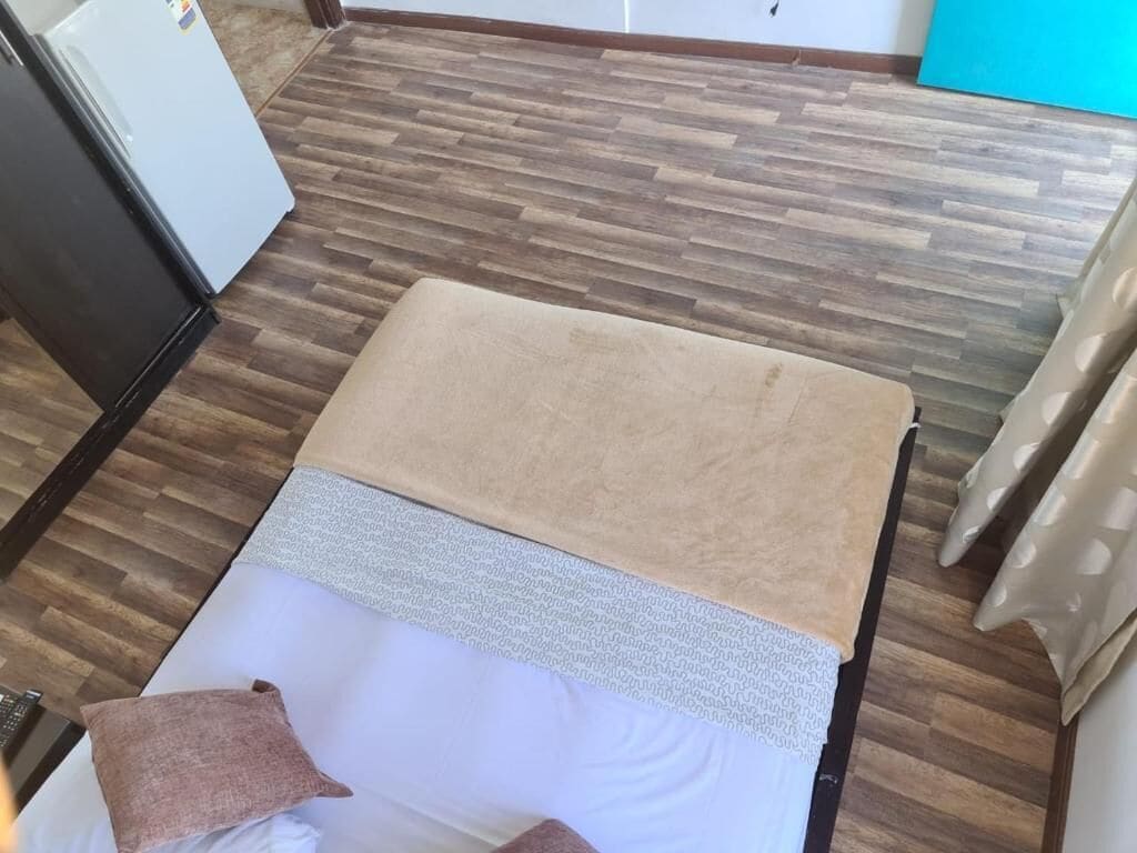 Una habitación con piso de madera, una cama con una manta color beige, un almohadón blanco y un almohadón marrón.