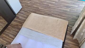 Una habitación con piso de madera, una cama con una manta color beige, un almohadón blanco y un almohadón marrón.