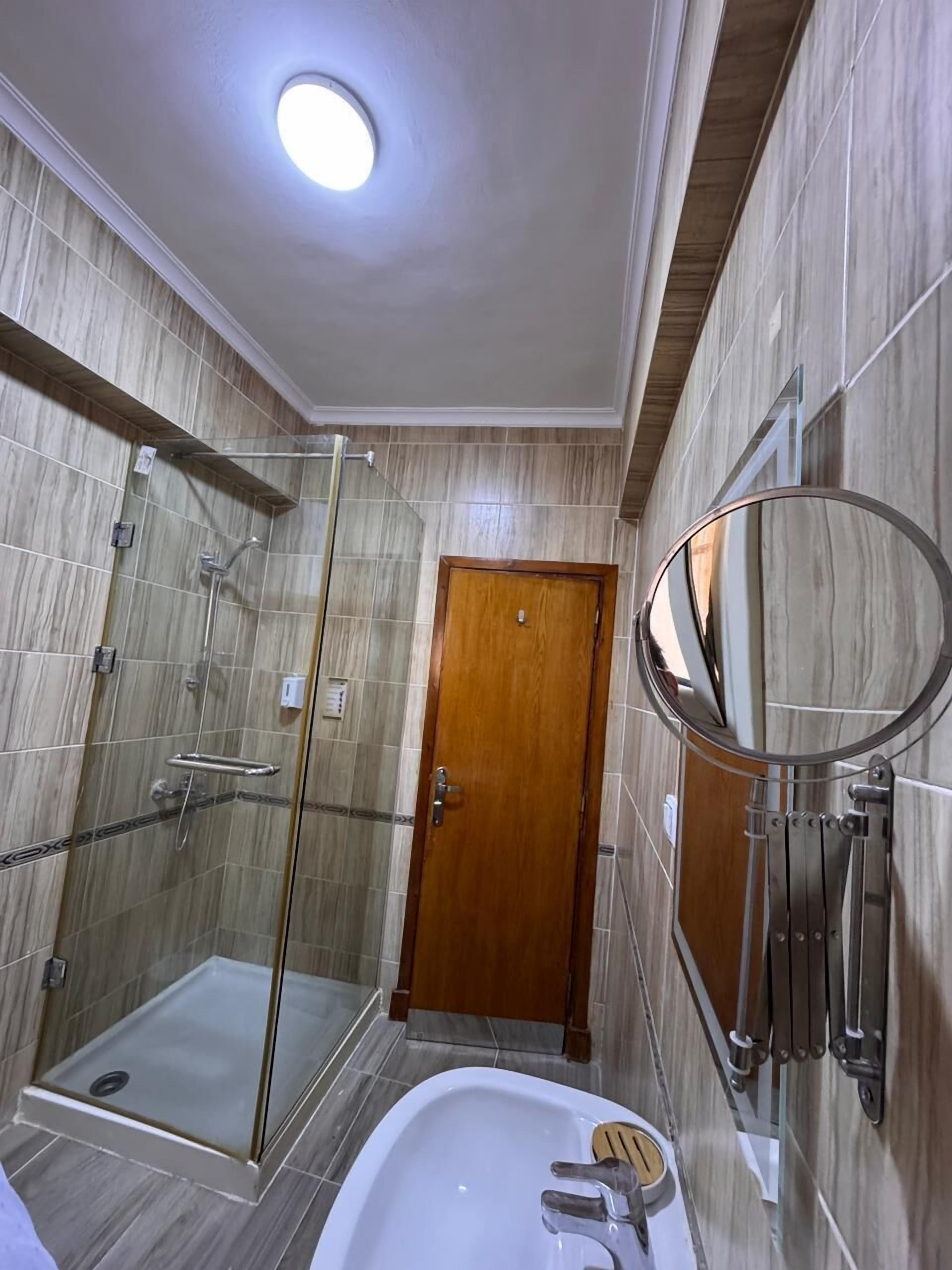 A modern bathroom with a glass shower enclosure, a wooden door, a white sink, and a round mirror.