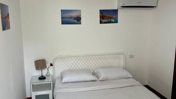 Un dormitorio con cama, mesitas de noche blancas, una lámpara y dos unidades de aire acondicionado instaladas en la pared.