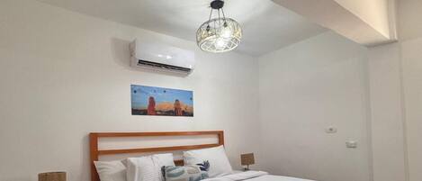 Un dormitorio con cama de madera, ropa de cama blanca, aire acondicionado de pared, un cuadro y una lámpara moderna.