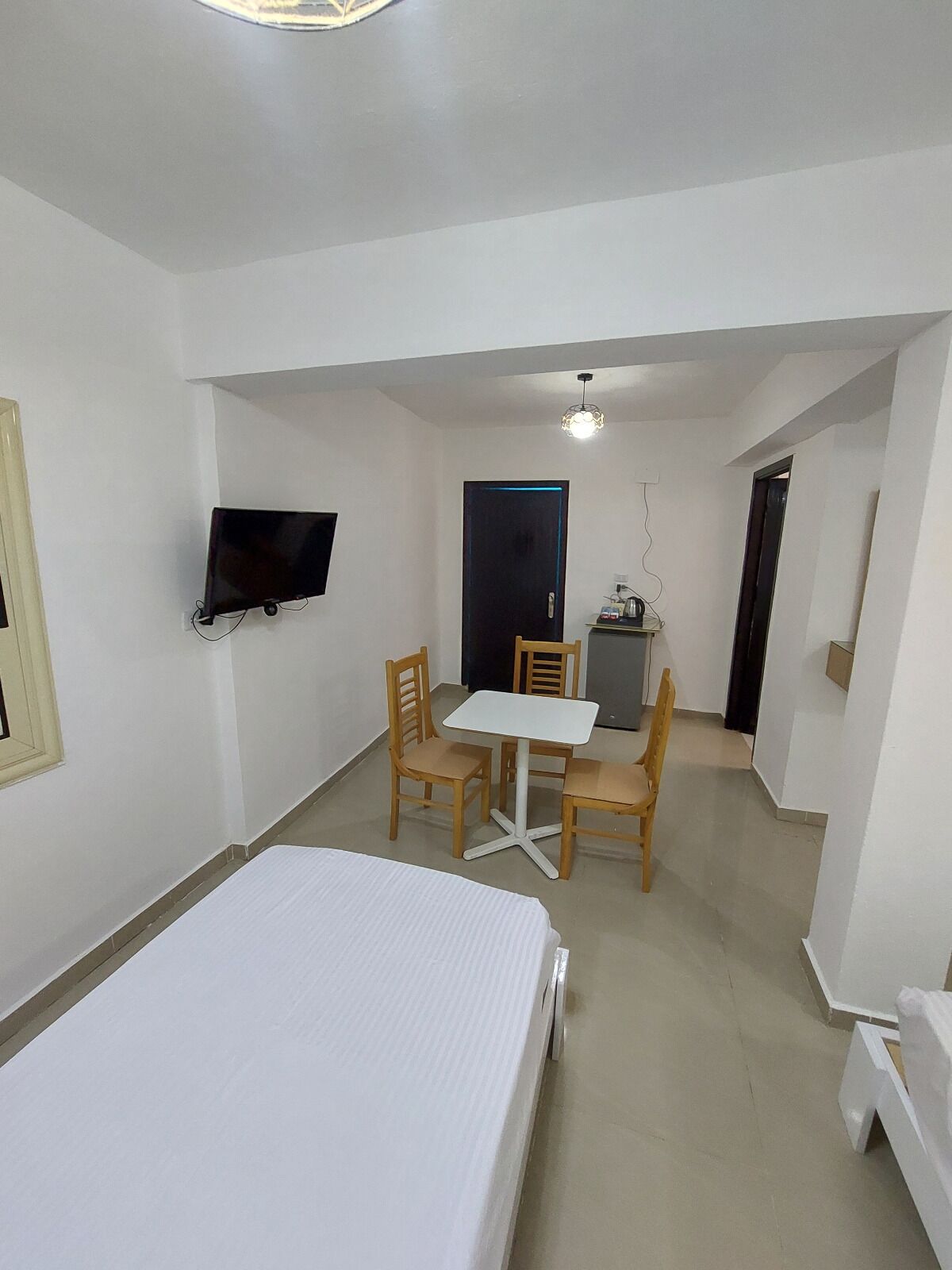 A room with a bed, a TV mounted on the wall, a table with chairs, and a door leading to another room.