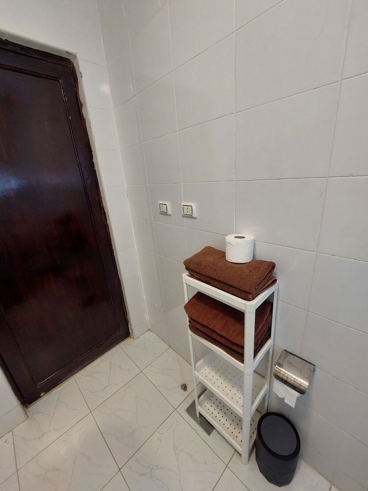 A bathroom with white tiled walls, a dark wooden door, a white shelf with towels, a toilet paper holder, and a trash bin.