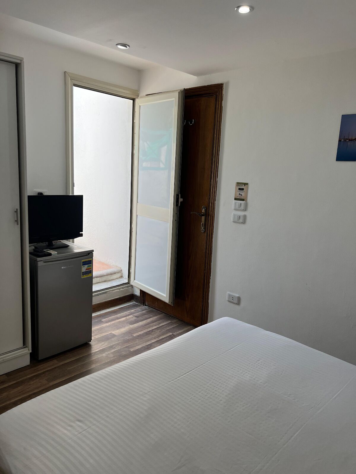 A small, clean room with a bed, a desk with a computer, a refrigerator, and a sliding glass door leading to a balcony.