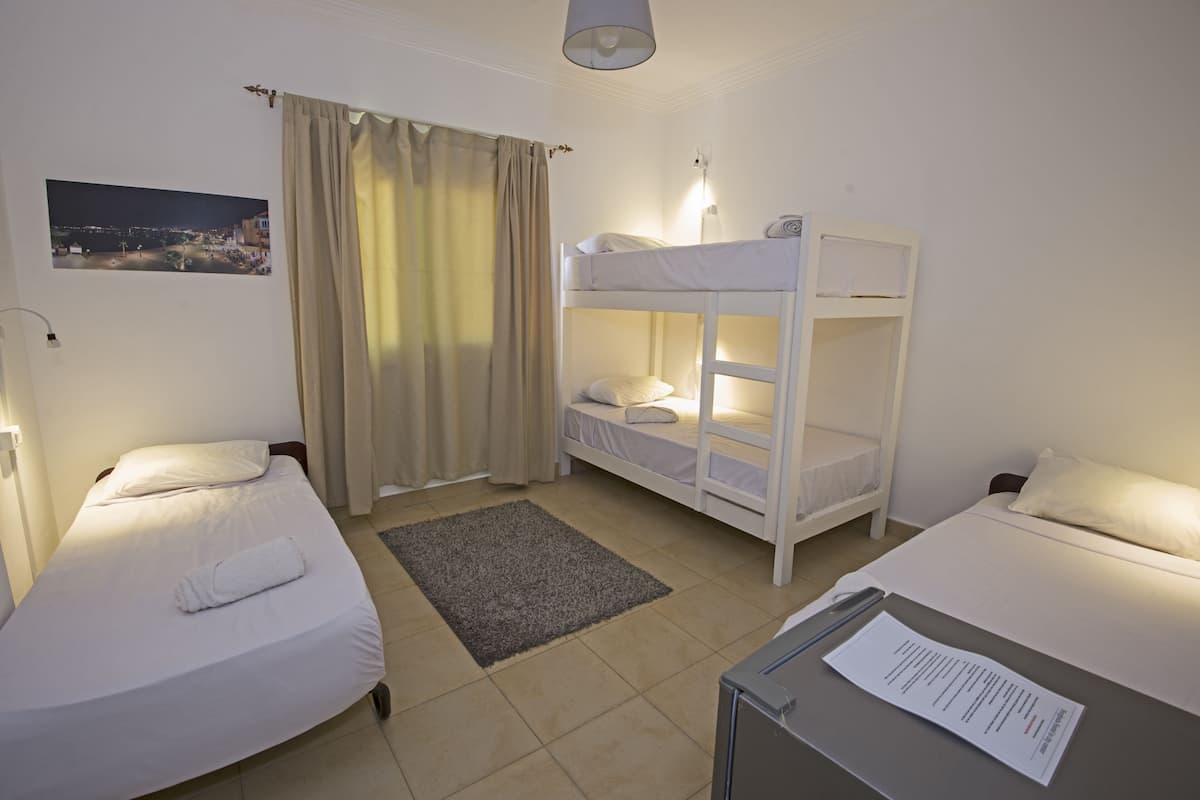 A hotel room with two single beds, a bunk bed, a grey rug, a night view photo on the wall, and a desk with a paper.