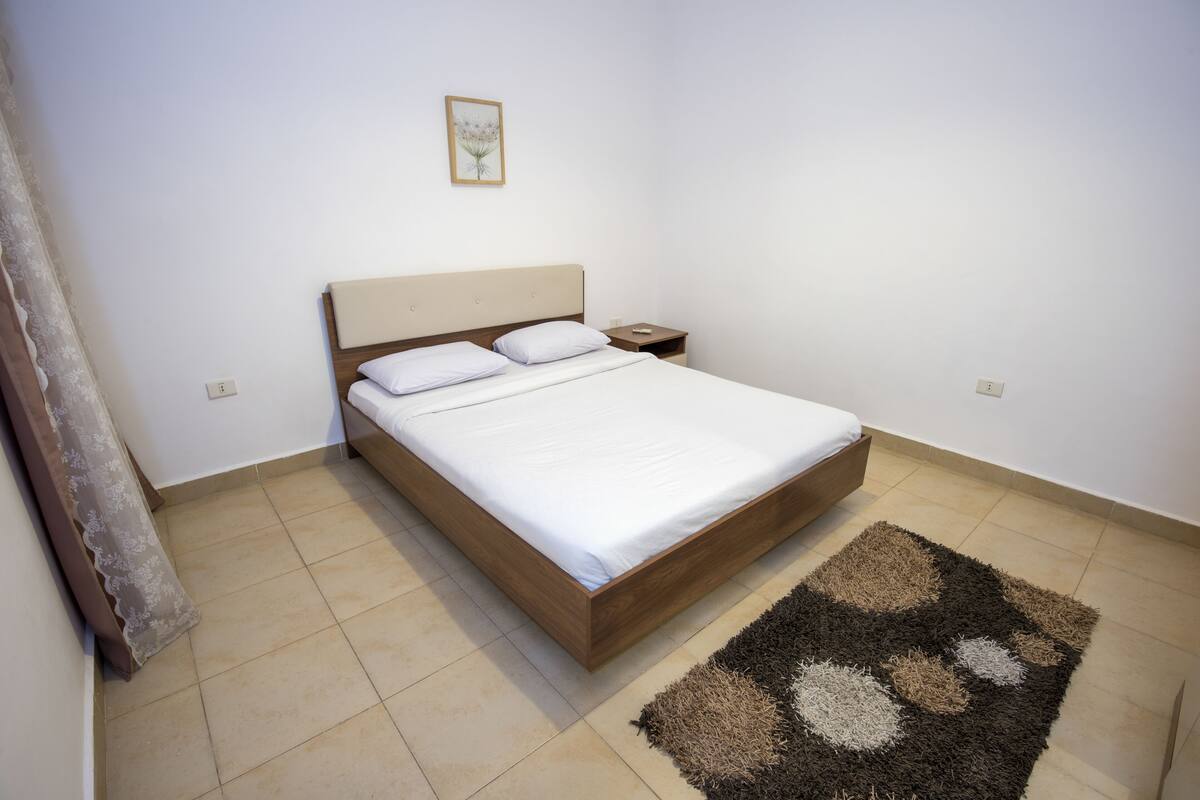 A single bed with a wooden frame and white bedding, a nightstand with a lamp, and a patterned rug on a tiled floor.