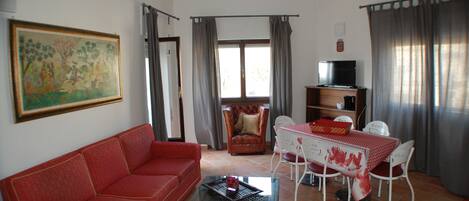 A living room with a red sofa, a glass coffee table, a dining table with chairs, a television, and a painting on the wall.