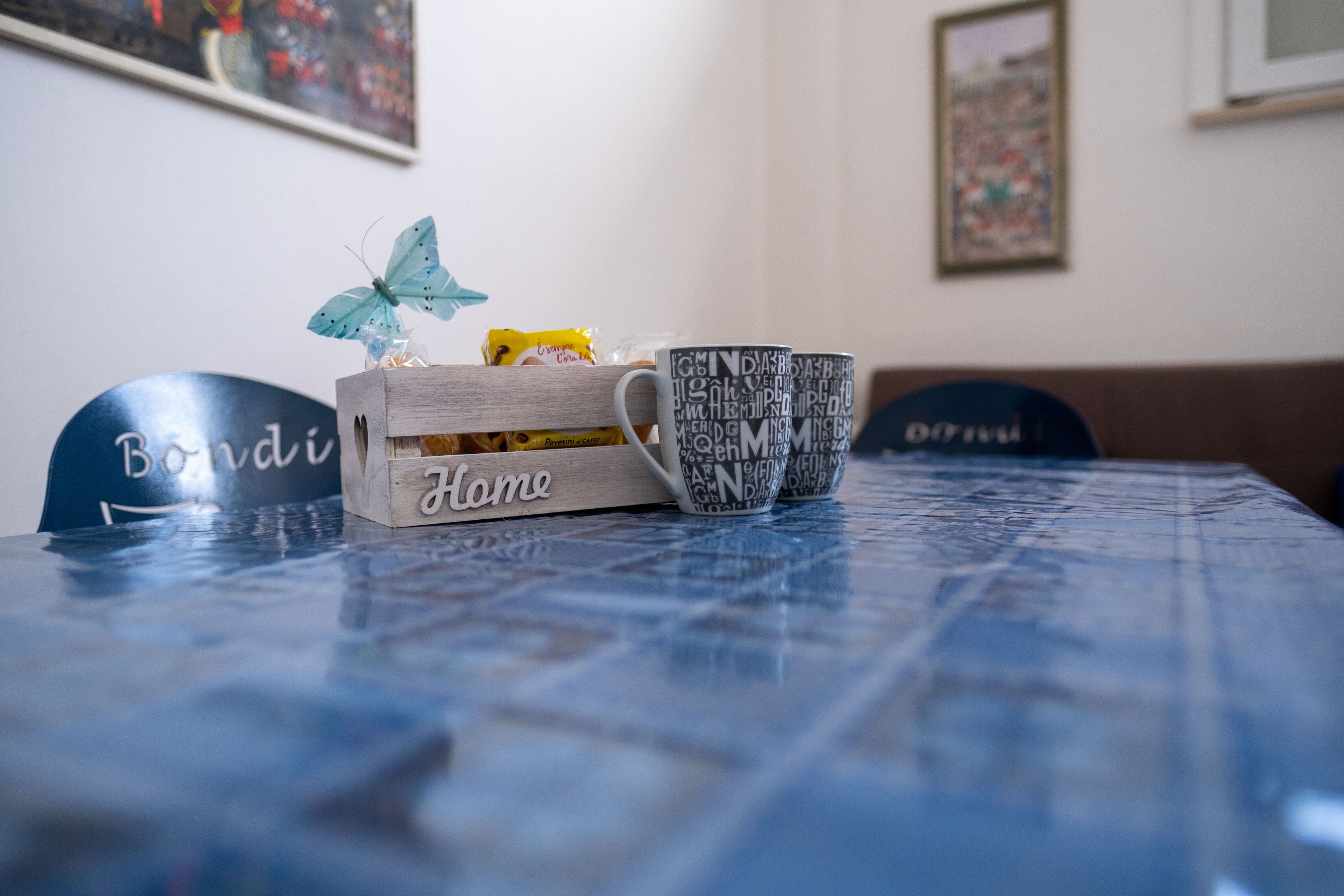 Una mesa con mantel azul, una caja de madera con la etiqueta 'Home', una taza con un diseño de palabras y una decoración de mariposa.