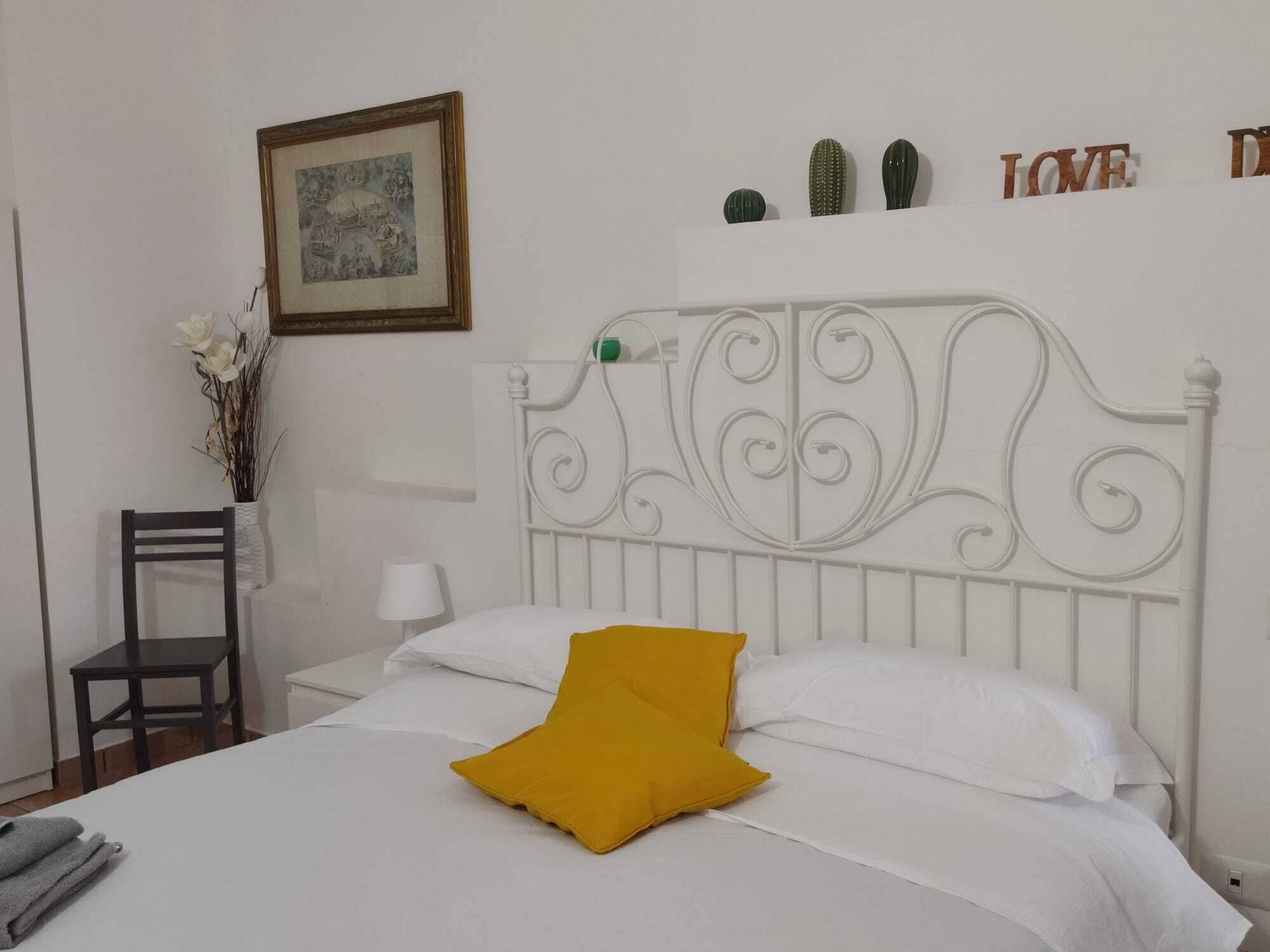 Un lit bien fait, avec du linge de lit blanc et un seul coussin jaune moutarde, un tableau encadré au mur, une petite collection de cactus et une chaise sur laquelle est posé un vase de fleurs.