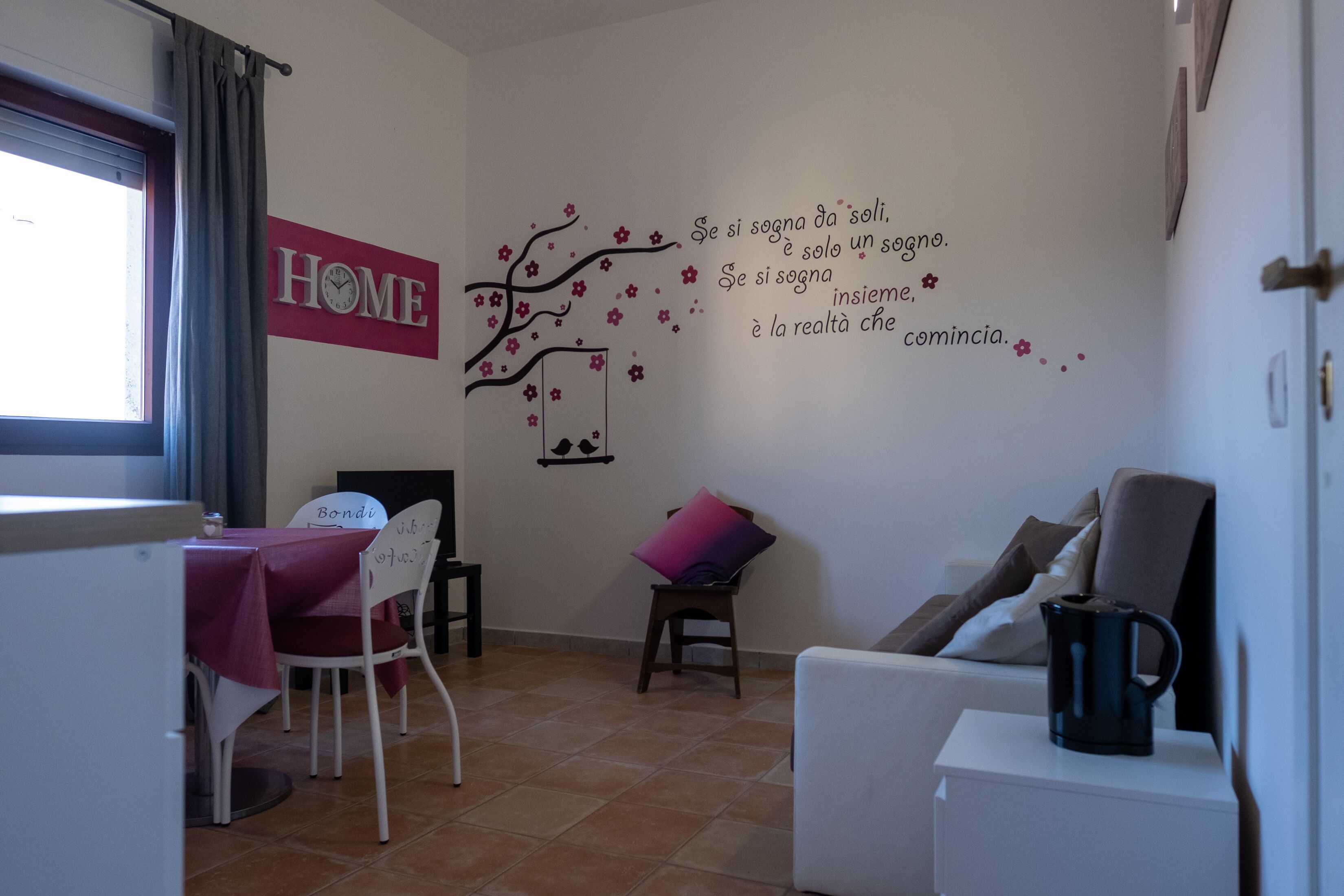 Habitació acollidora amb zona de menjador, un sofà i una paret amb un rètol que diu 'HOME' i un arbre decoratiu amb flors.
