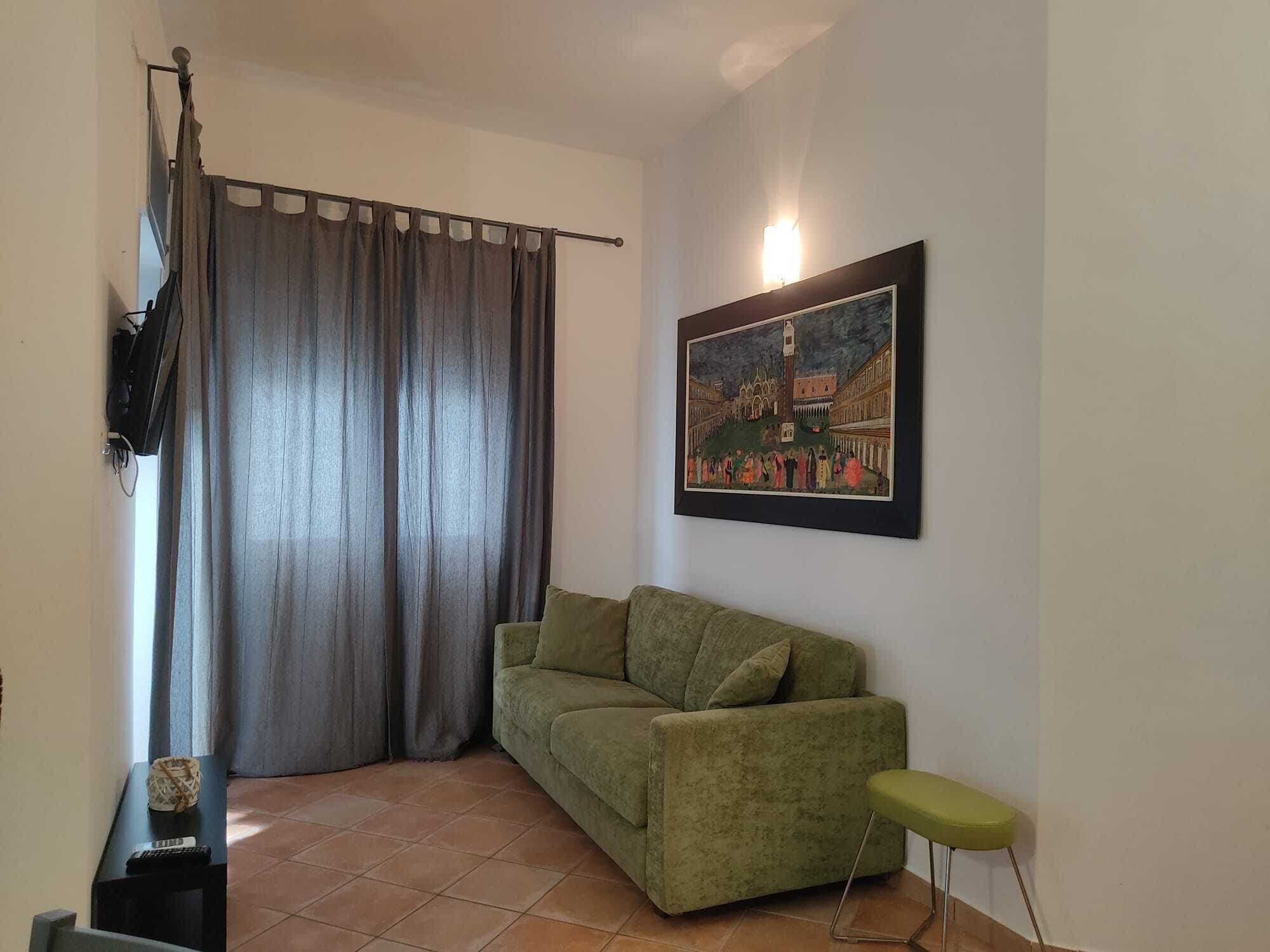 Un salón con un sofá verde, una mesita con un control remoto, un cuadro en la pared y una cortina.