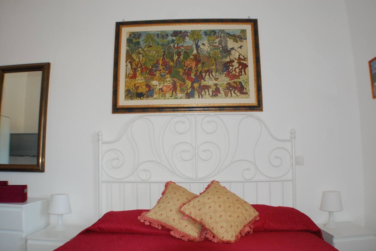 A bedroom with a white metal headboard, a red bedspread, two pillows, a framed painting, and a mirror on the wall.