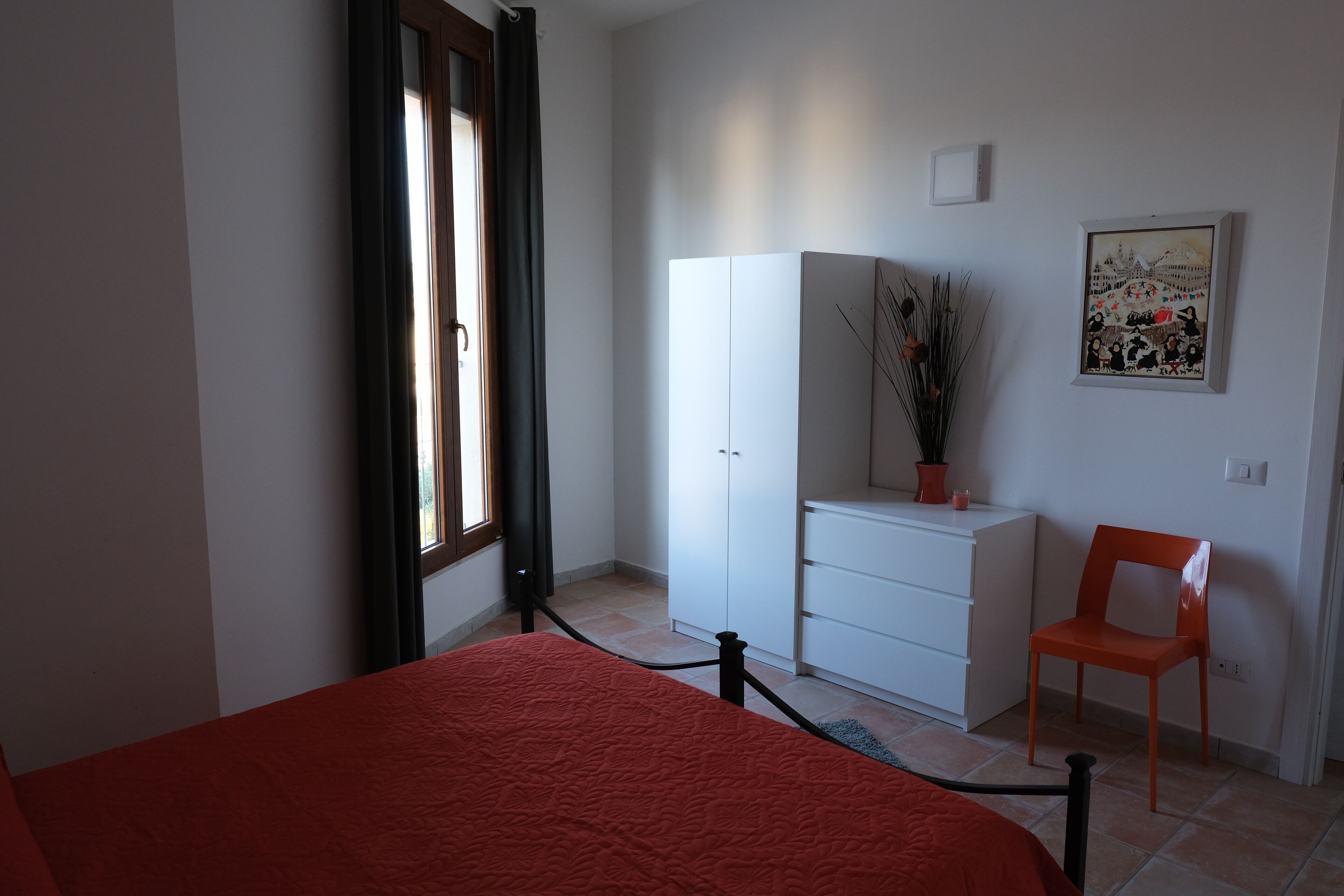 A bedroom with a bed, a white wardrobe, a chair, and a painting on the wall.