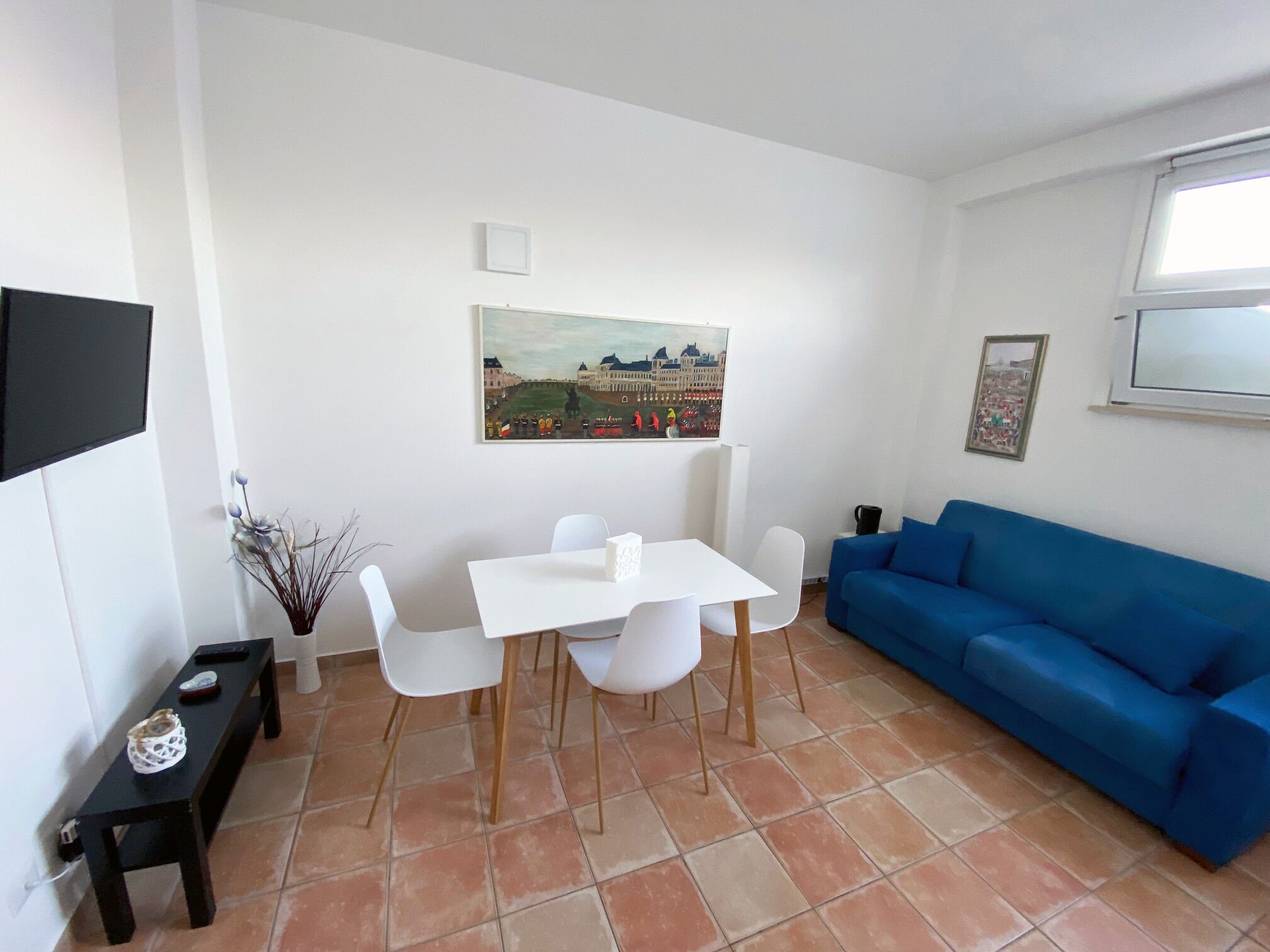 A living room with a white table, four white chairs, a blue sofa, a painting on the wall, and a window.