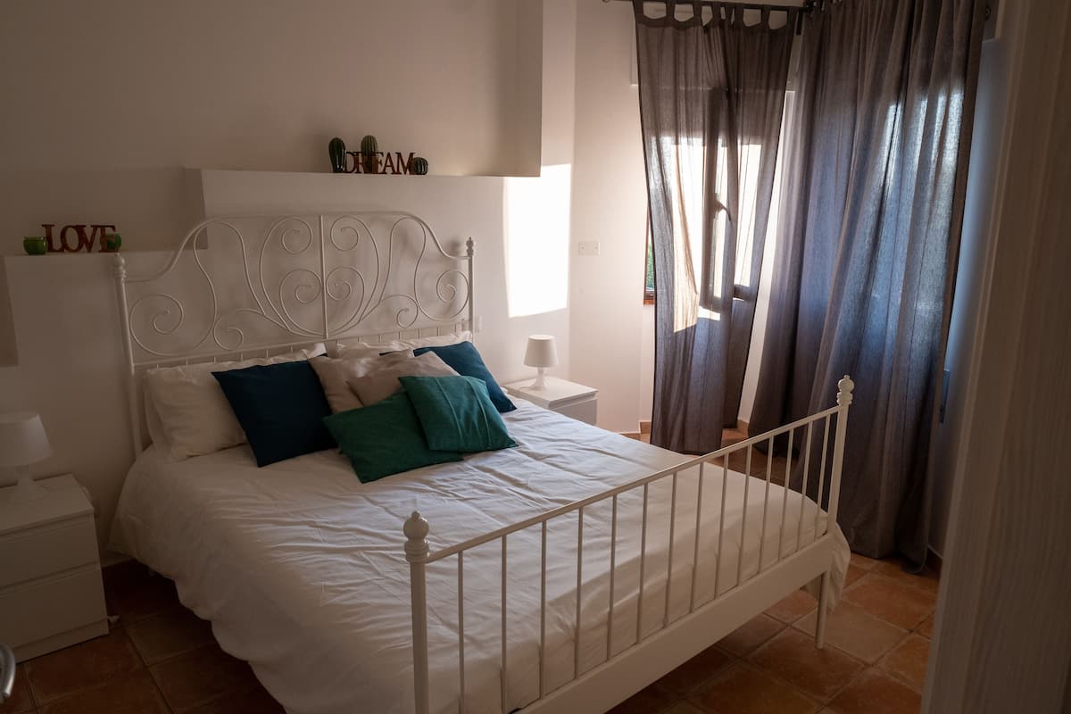 A neatly made bed with white bedding and teal pillows, a white nightstand with a lamp, and a window with curtains.