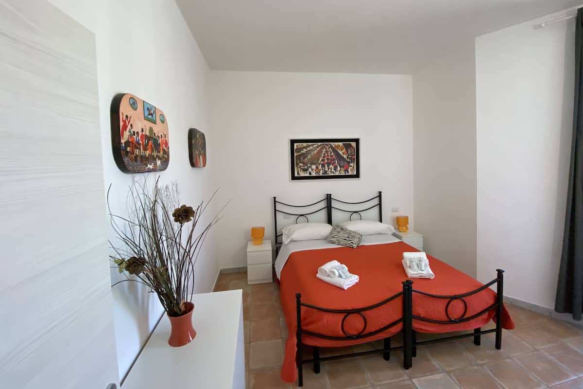 A bedroom with a bed, a nightstand, a bedside table, a potted plant, and wall art.
