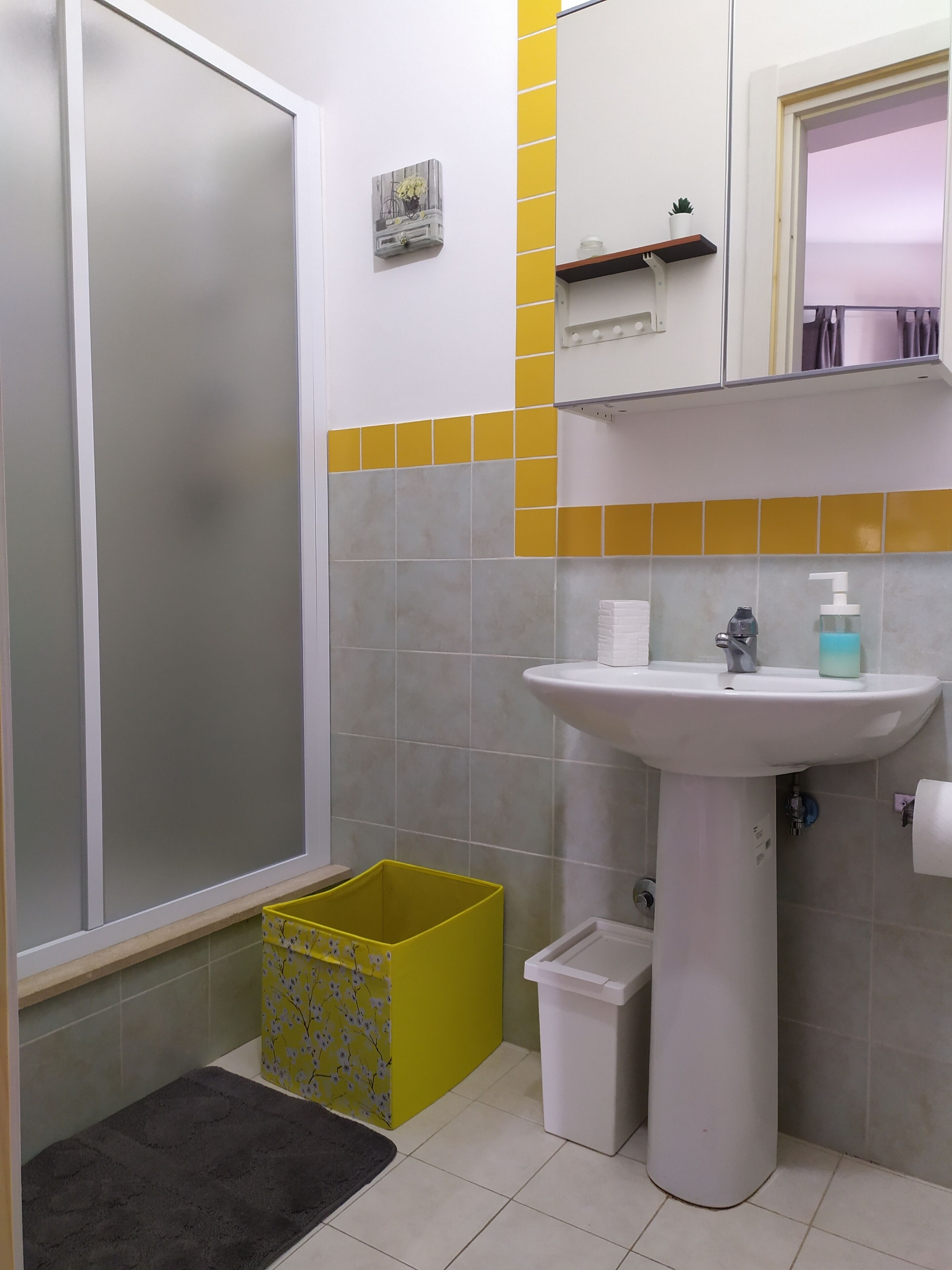 Une salle de bain avec une douche, un lavabo muni d’un distributeur de savon, une poubelle et un panier jaune.