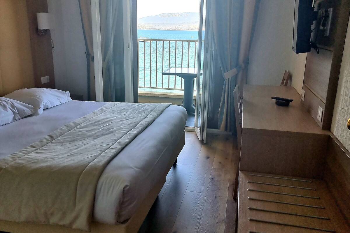 A hotel room with a bed, a desk, a chair, a TV, and a balcony with a view of the sea.