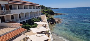 Ein Hotel mit mehreren Balkonen mit Blick auf das klare, blaue Meer, Sitzgelegenheiten im Freien mit Sonnenschirmen und einer felsigen Küstenlinie.