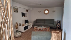 Sala de estar con un sofá, una mesa de centro y un televisor sobre un soporte blanco. Hay un jarrón decorativo y una cesta tejida en el suelo.