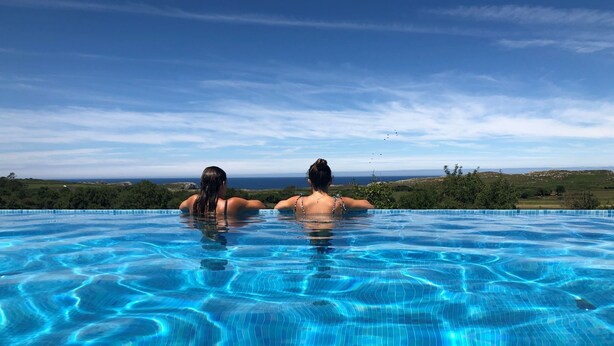 Ein klarer, blauer Infinity-Pool mit zwei entspannten Personen am Rand und Blick auf eine reizvolle Landschaft.